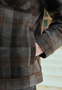 Persona che indossa una giacca di lana a scacchi marrone e grigia con una mano infilata nella tasca laterale, in piedi contro uno sfondo di legno.