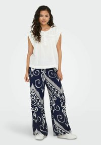 Blouse blanche sans manches avec détails de boutons associée à un pantalon large marine présentant un motif blanc tourbillonnant. Le mannequin se tient avec assurance.