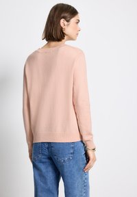 Femme avec des cheveux bruns courts portant un pull rose clair et un jean bleu, de profil contre un fond blanc uni.