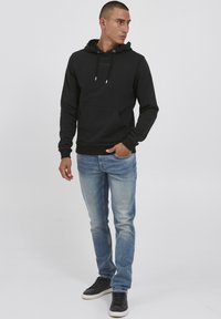 Svart hettegenser med frontlomme, snøring i hetta og ribbestrikkede mansjetter, kombinert med lyseblå slitte jeans og svarte joggesko.