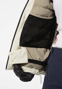 Gewatteerde jas met een beige binnenzijde, zwart mesh zakje en drukknoopsluiting. Voorzien van een trekkoord aan de onderrand en een zwarte handschoen.