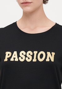 Svart t-skjorte med rund hals som har fete, glitrende gullbokstaver som staver "PASSION" over brystet. Mykt bomullsmateriale.