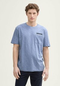 Camiseta azul de manga corta con bolsillo, que presenta una tela texturizada y un cuello redondo, llevada por un modelo contra un fondo neutro.