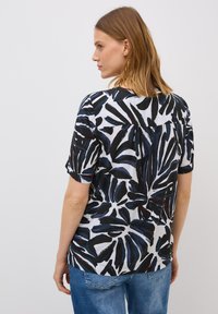 Eine Frau mit langen braunen Haaren ist von der Seite zu sehen, trägt ein gemustertes Oberteil mit schwarzen, blauen und weißen Blattmustern sowie eine blaue Jeans.