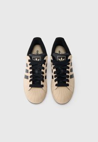 Béžové tenisky s rebrovanou špičkou, čiernymi akcentmi a tromi čiernymi pruhmi. Majú čierne šnúrky a logo na jazyku.