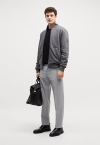 Giacca bomber grigia, t-shirt nera, pantaloni grigi chiari e scarpe nere. Tiene una borsa nera con un design strutturato e accenti metallici.