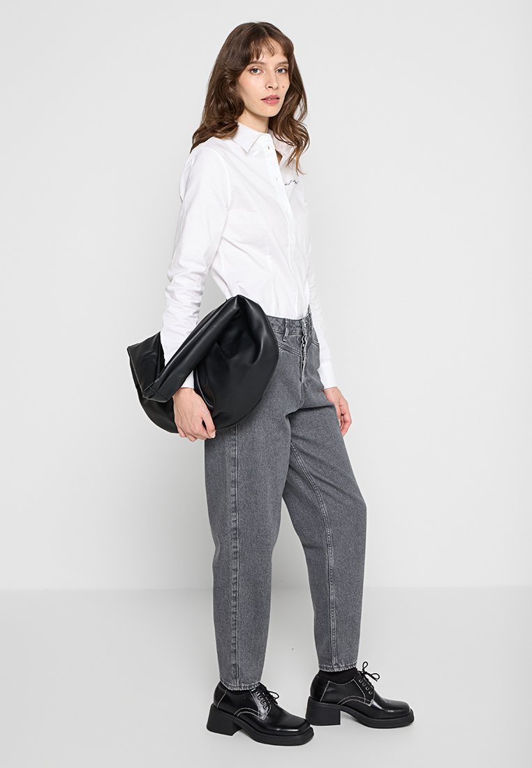 Borsa in pelle nera morbida con design piegato, indossata da una persona in camicia bianca e jeans grigi a gamba stretta, in piedi su uno sfondo neutro.
