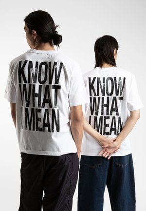 Hvit bomulls t-skjorter med svart tekst "KNOW WHAT I MEAN" på ryggen, båret av to individer, kombinert med svarte og blå bukser.
