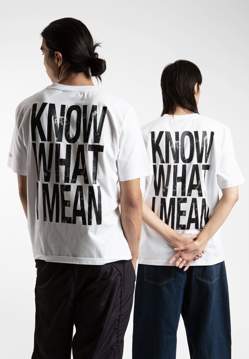 White cotton t-shirts with black text "KNOW WHAT I MEAN" on the back, worn by two individuals, paired with black and blue pants.