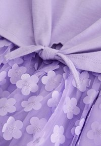 Vêtement violet avec un haut uni à détail noué et un bas à couches de tissu transparent orné de motifs floraux et d'accents métalliques.