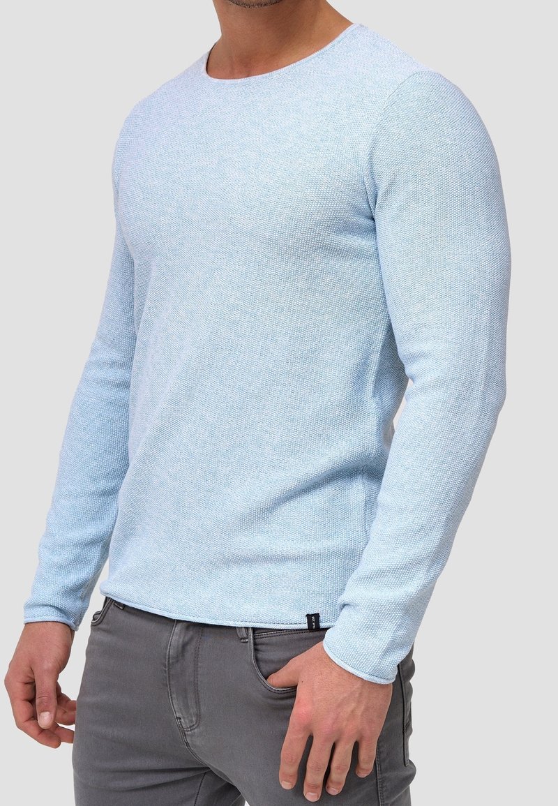 Pull over en bleu clair à manches longues avec un tissu texturé, design à col rond et une petite étiquette de marque à l'ourlet, associé à un pantalon gris.
