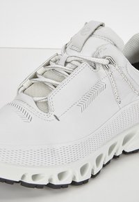 Weiße Sportschuh mit einem strukturierten Leder-Obermaterial, Netzakzenten und einer klobigen Sohle, die Aussparungen für Belüftung und Dämpfung aufweist.