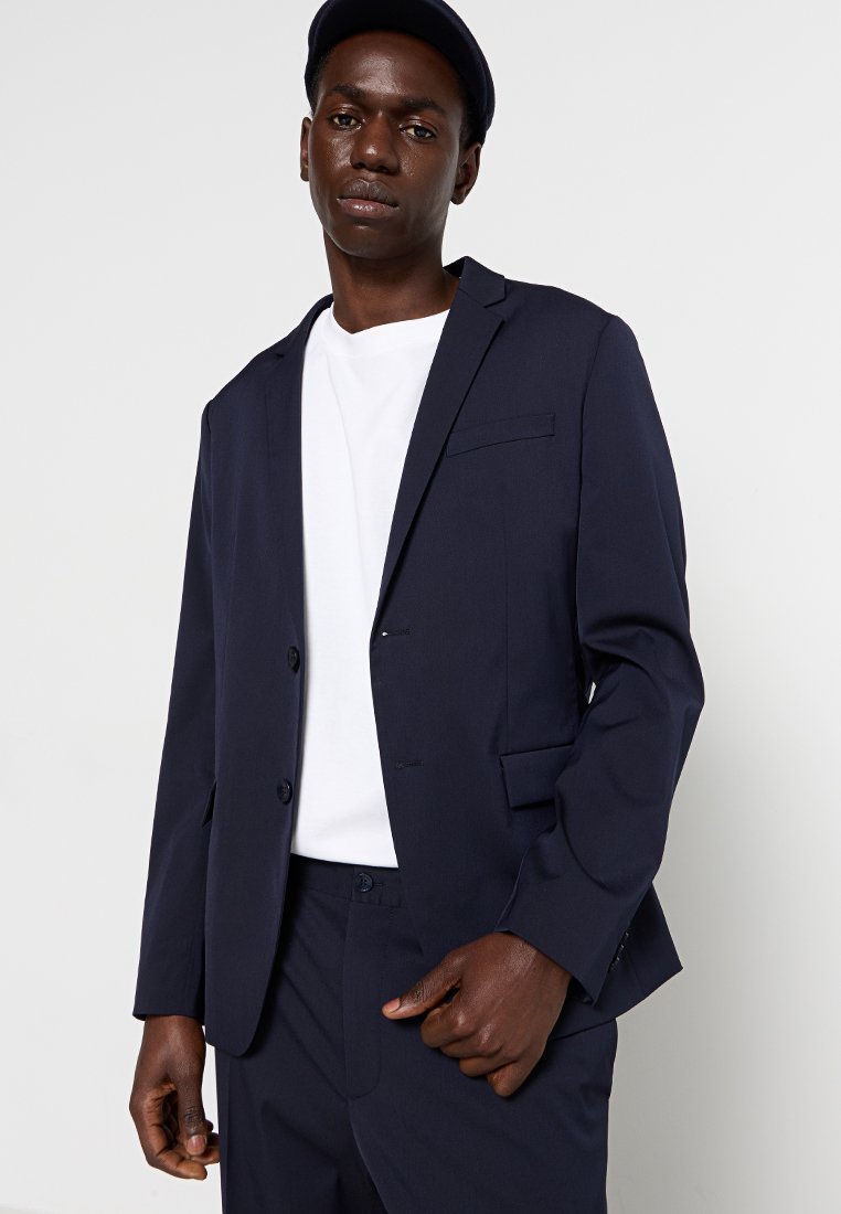 Blazer marine en tissu lisse, doté d'une fermeture à un bouton, de revers crantés et d'une poche poitrine. Porté sur un T-shirt blanc.