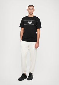 Melns kokvilnas T-krekls ar īsām piedurknēm, centrā balts apļveida logotips, kombinēts ar brīva piegriezuma krēmkrāsas biksēm un melniem sporta apaviem.