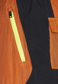 Oranžová a černá látka bundy s mřížkovaným vzorem, která má výrazný žlutý zip a boční kapsu s kontrastním akcentem.