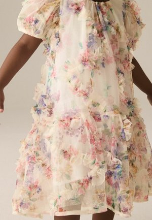 Enfant portant une robe transparente à motif floral avec des bords volantés et des manches courtes bouffantes sur un fond uni.