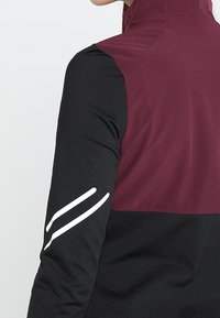 Czarna i burgundowa sportowa kurtka z wysokim kołnierzem. Ma białe paski akcentujące na rękawie. Teksturowany materiał, dopasowany krój.