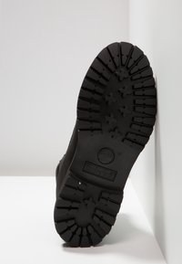 Semelle noire robuste d'une botte Timberland montrant des crampons profonds et le logo, debout contre un coin blanc de mur.