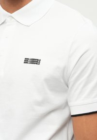 Fehér, rövid ujjú póló, a bal mellen háromszor ismétlődő fekete "KARL LAGERFELD" logóval.