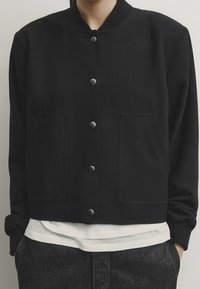 Personne portant une veste noire boutonnée avec deux poches frontales, un sous-maillot blanc et un pantalon en denim foncé, les mains dans les poches.