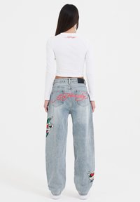 Hvid langærmet crop top med et rødt logo på ryggen, parret med lyseblå baggy jeans med farverige broderede designs.