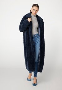 Mørkeblå lang pelsfrakke med en tekstureret, blød overflade. Båret over en grå rullekrave og blå jeans, med mønstret foret og blå sko.