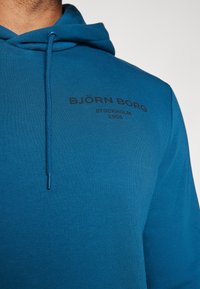 Modrá mikina s kapucňou vyrobená z textúrovanej tkaniny. Obsahuje kapucňu s šnúrkou a čierny potlačený text "BJÖRN BORG STOCKHOLM 1986."