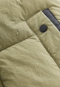 Chaqueta impermeable de color verde oliva con una superficie texturizada, que presenta gotitas de agua, secciones acolchadas y un acento de botón negro.