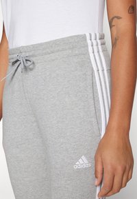 Sivé tepláky vyrobené z bavlnenej zmesi s elastickým pásom na šnúrku a tromi bielymi pruhmi po strane, biele logo Adidas.