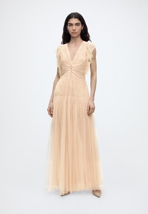 Žena s dlhými tmavými vlasmi, oblečená v dlhých broskyňových tylových šatách s nariasenými krátkymi rukávmi a hlbokým výstrihom do V, stojaca na jednoduchom pozadí.