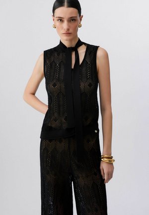 Femme aux cheveux coiffés en arrière porte un haut sans manches en dentelle noire avec cravate et un pantalon large assorti, accessoirisés de boucles d'oreilles et bracelets dorés.