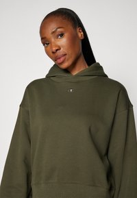 Olivgrüner Hoodie aus weichem Stoff, mit einer Fronttasche und einem kleinen gestickten Logo auf der Brust. Klassischer, lockerer Schnitt.