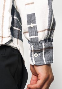 Camicia di flanella a quadri con tonalità navy, bianco e marrone, con polsini a bottone e tessuto testurizzato. Mostrata su uno sfondo di pantaloni neri.
