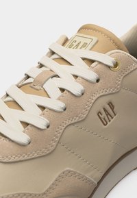 Zbliżenie na beżowy i brązowy sneaker z białymi sznurowadłami, panelami ze skóry i tkaniny oraz logo "GAP" na języku i boku.