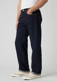 Jeans de denim azul oscuro con corte recto, que presentan herrajes de cobre, bolsillos frontales y costuras amarillas sutiles, combinados con zapatillas blancas.