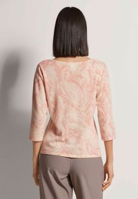 Leichtes, rosa gemustertes Oberteil mit Dreiviertelärmeln und rundem Halsausschnitt, kombiniert mit hellgrauen Hosen. Weicher, strukturierter Stoff.