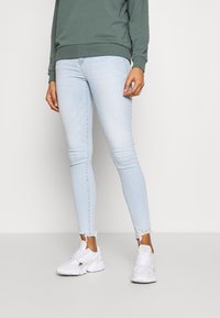 Pantalones vaqueros ajustados azul claro con dobladillo deshilachado, combinados con una sudadera verde y zapatillas blancas. El material parece ser mezclilla, con un diseño entallado.