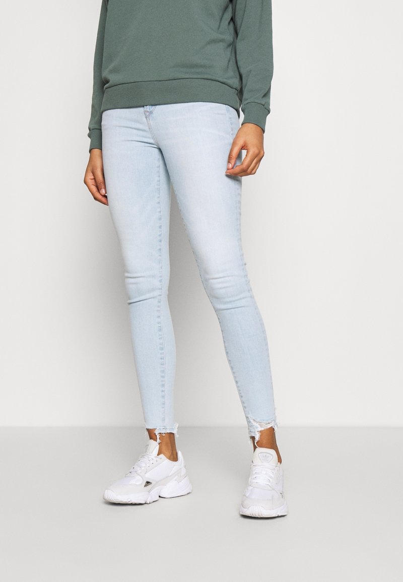 Pantalones vaqueros ajustados azul claro con dobladillo deshilachado, combinados con una sudadera verde y zapatillas blancas. El material parece ser mezclilla, con un diseño entallado.