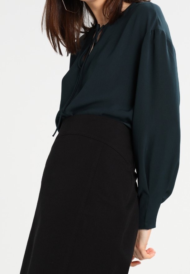 Femme portant un chemisier vert foncé à manches longues avec un lien au col et une jupe noire taille haute, debout devant un fond uni.