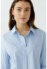 Blue striped blouse featuring vertical white stripes, embellished with silver sequins, and a classic collar design. Smooth fabric texture.