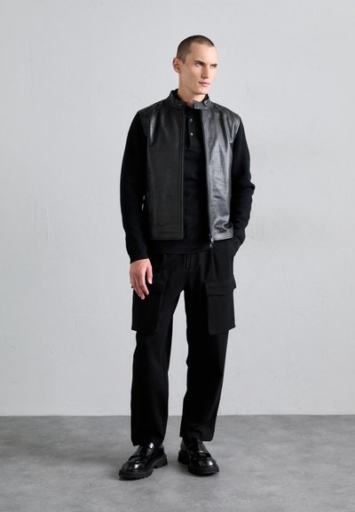 Black leather vest with zip closure, paired with a black long-sleeve shirt and loose-fit black cargo pants. Black chunky shoes complete the look.