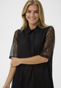 Smaidoša sieviete ar blondiem matiem valkā melnu mežģīņu kleitu ar caurspīdīgām mežģīņu piedurknēm un pogāmu apkakli, pozējot pret vienkāršu fonu.