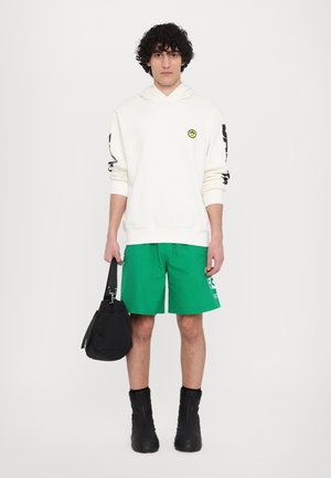 Kremfarget hettegenser med en gul smiley-merke, grønne shorts og svarte glidelåsstøvler. Holder en svart bag med en stropp.