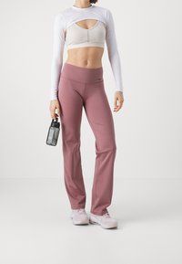 Completo sportivo composto da una maglietta bianca a maniche lunghe, reggiseno beige e pantaloni rosa a vita alta; tiene in mano una bottiglia d'acqua scura.
