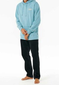 Jasnoniebieska bluza z kapturem wykonana z miękkiej tkaniny, z kieszenią z przodu, sznurkami i logo; w połączeniu z czarnymi spodniami. Bez obuwia.