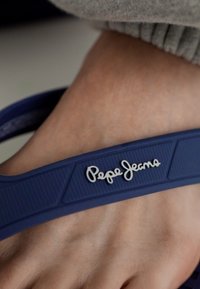Tummansininen flip-flop, jossa on tekstuurilla varustettu hiho, jossa on kohotettuja yksityiskohtia ja valkoinen "Pepe Jeans" -logo, lepäämässä paljaan jalan päällä.