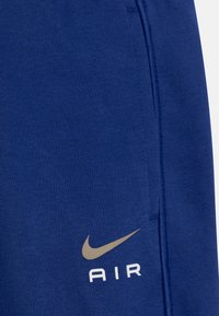 Pantalones cortos de algodón azul con un bolsillo lateral, con el logo del swoosh en dorado de Nike y el texto "AIR" en blanco. Texture suave y diseño casual.