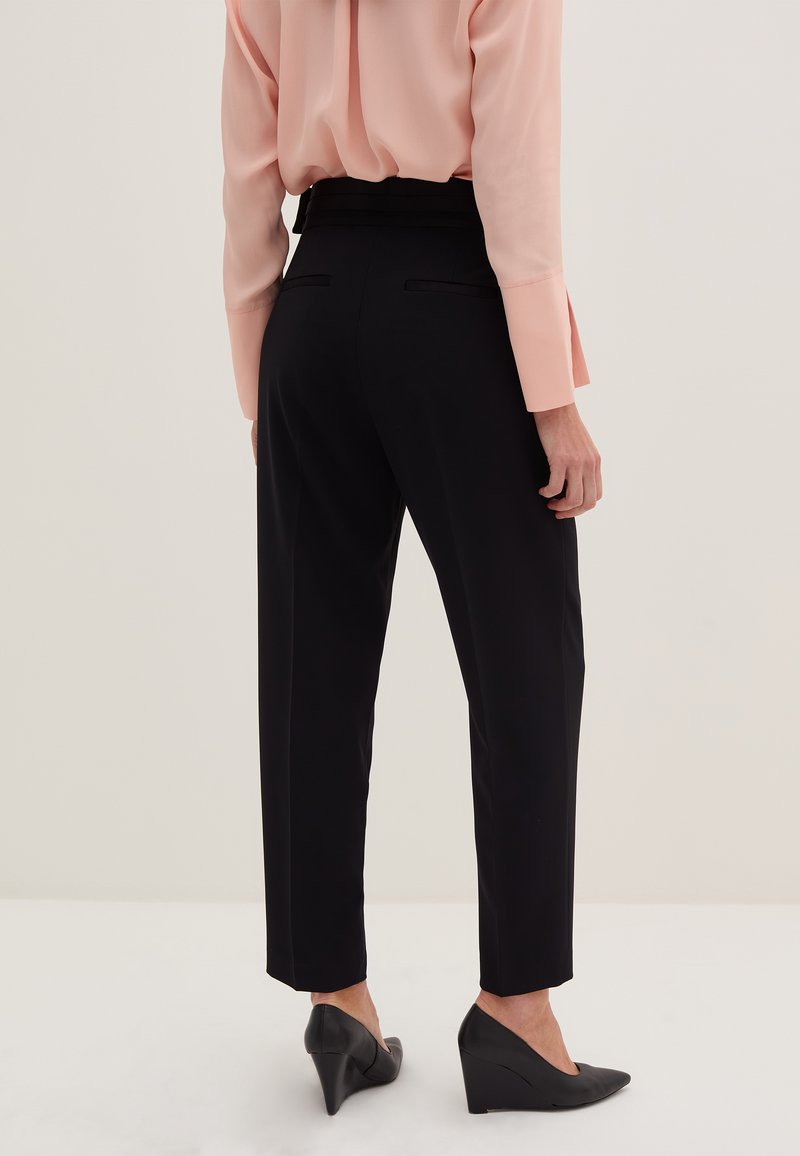Schwarze taillierte Hose mit hoher Taille, glattem Stoff und zwei Gesäßtaschen, kombiniert mit einer pfirsichfarbenen Bluse mit langen Ärmeln und schwarzen Keilabsätzen.