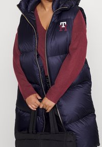 Marineblauer Pufferweste mit Kapuze und goldener Reißverschluss, kombiniert mit einem roten Strickpullover. Das Model hält eine schwarze Tragetasche. Das „TH“-Logo ist auf der Weste zu sehen.