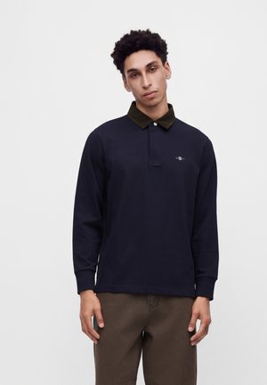 Ein junger Mann mit kurzen lockigen Haaren trägt ein marineblaues Langarm-Polohemd mit dunklem Kragen und braune Hose vor einem schlichten weißen Hintergrund.